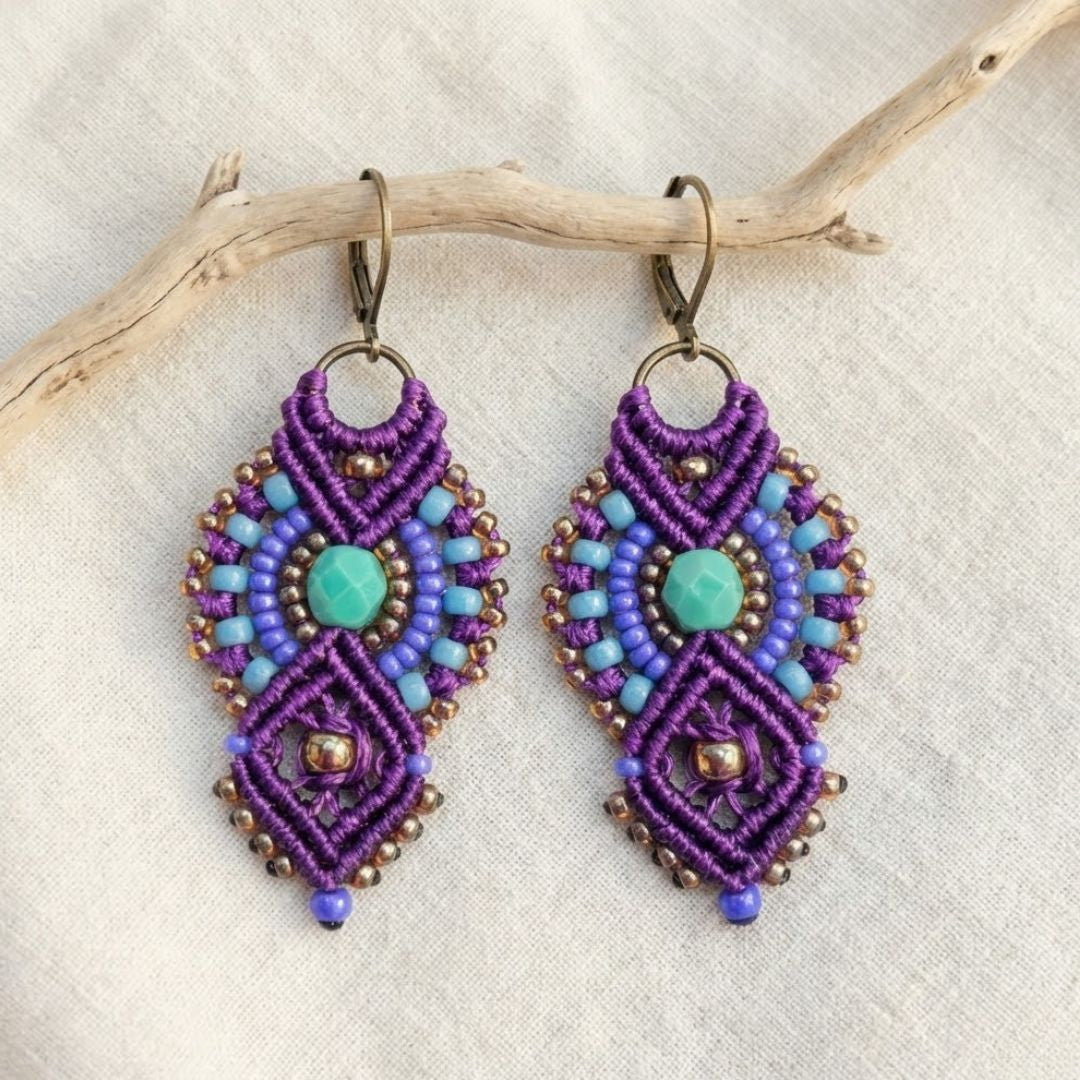 עגילי מקרמה אינקה סגול וטורקיז עם חרוזי זכוכית תלויים על ענף מעל בד פשתן