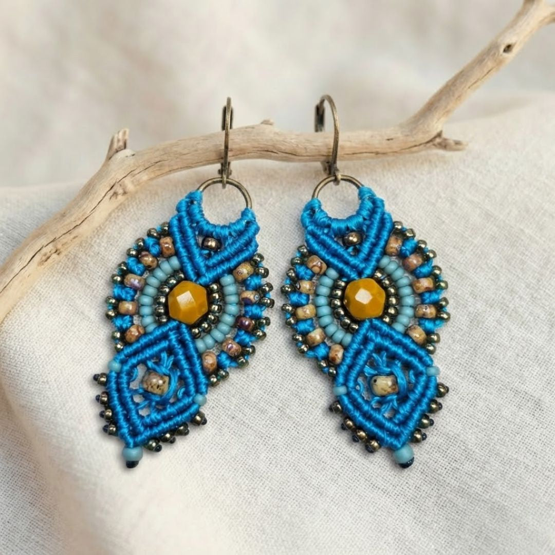 עגילי מקרמה אינקה טורקיז וחרדל עם חרוזי זכוכית תלויים על ענף מעל בד פשתן