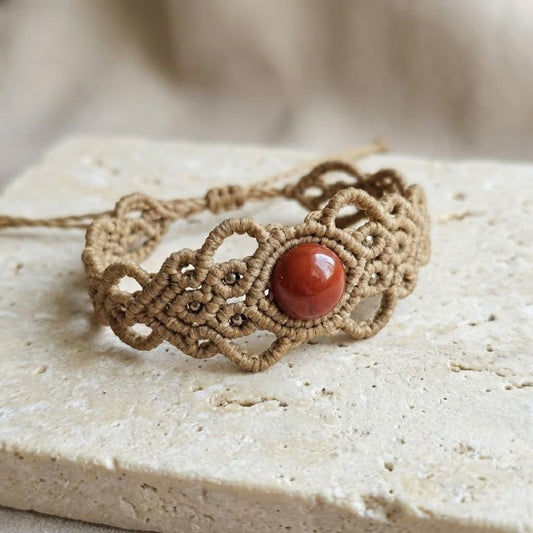 צמיד 'גאיה' חום קאמל עם ג'ספר אדום וחרוזי בראס על משטח אבן בהיר, מבנה הסריגה מודגש. סגירה מתכווננת נראית בפריים