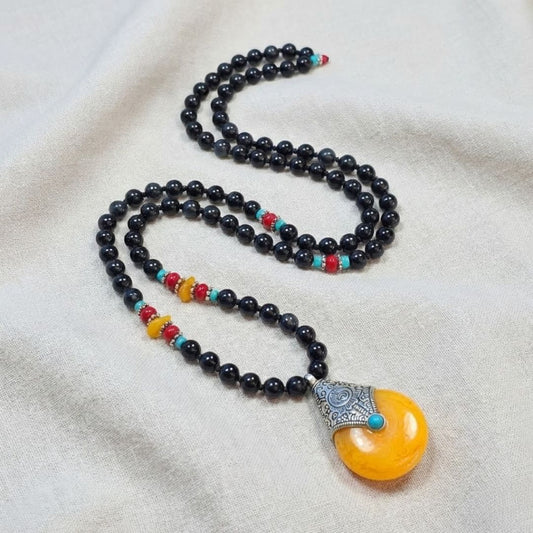 שרשרת מאלה 'עין האש' עם אבני אובסדיאן ותליון ענבר טיבטי בשילוב חרוזי ענבר, אדום וטורקיז מונחת על בד טבעי