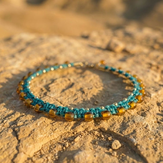 צמיד רגל מקרמה 'גואה' טורקיז עם חרוזי זכוכית ענבריים וחרוזי בראס מונח על אבן מדברית באור שקיעה