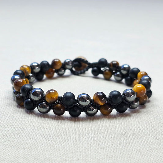 צמיד מקרמה עם אבני עין הנמר, המטייט ואוניקס מט מונח על בד בגוון טבעי, תקריב סריגת החוט ושילוב האבנים