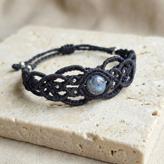 צמיד 'גאיה' שחור עם לרוויקייט וחרוזי בראס על משטח אבן בהיר, דגש על סריגת חוט שעווה