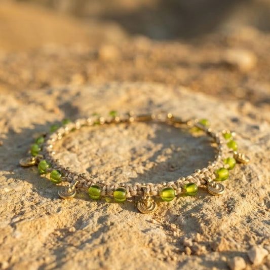 צמיד רגל 'גואה' בז' טבעי עם חרוזי זכוכית ירוקים ותליוני בראס עגולים מונח על משטח אבן מדברית