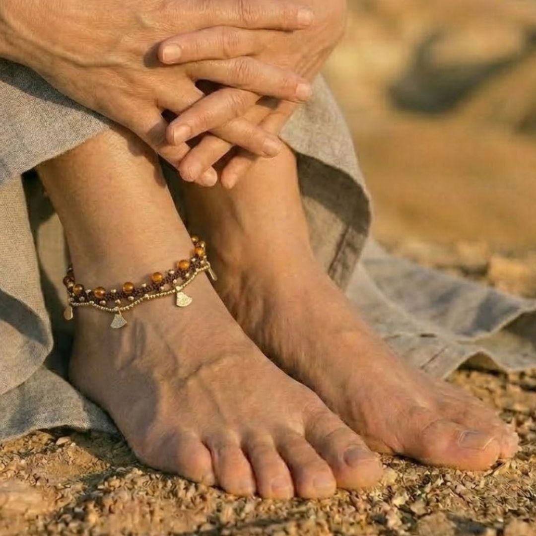 צמיד רגל פושקר מקרמה חום עם חרוזי זכוכית ענבריים וחרוזי בראס על קרסול יחף בנוף מדברי