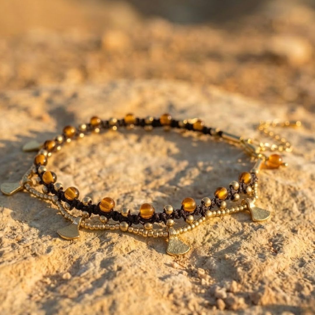 צמיד רגל פושקר חום בעבודת מקרמה עם חרוזי זכוכית ענבריים ואלמנטים מבראס מונח על אדמה מדברית