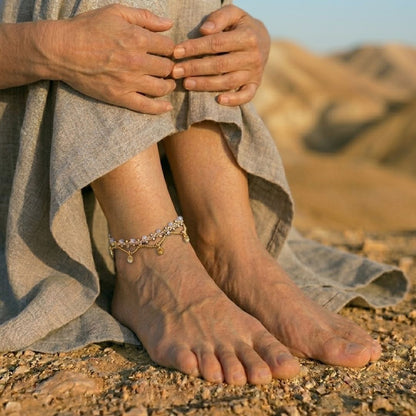 צמיד רגל פושקר מקרמה בהיר עם חרוזי זכוכית תכלת ותליוני בראס על קרסול בנוף מדברי